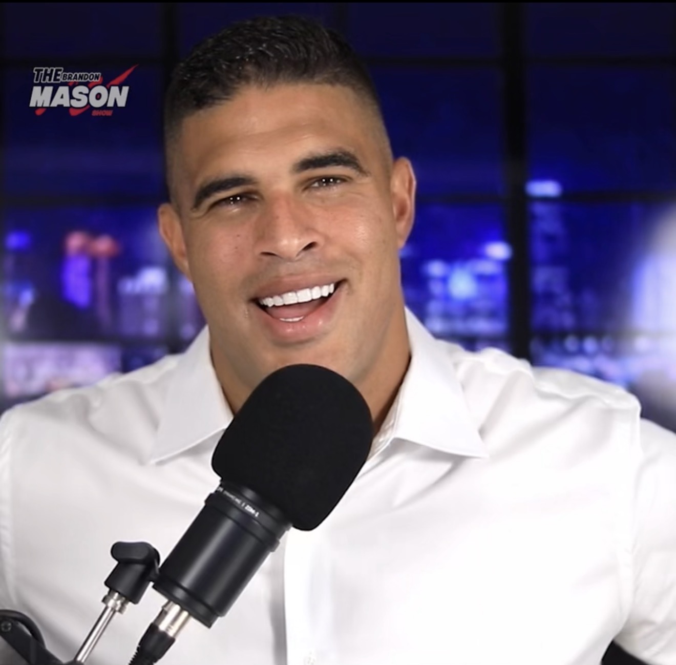 Brandon Mason, known as The Plug, wearing a white shirt at a black studio microphone with a purple cityscape behind him