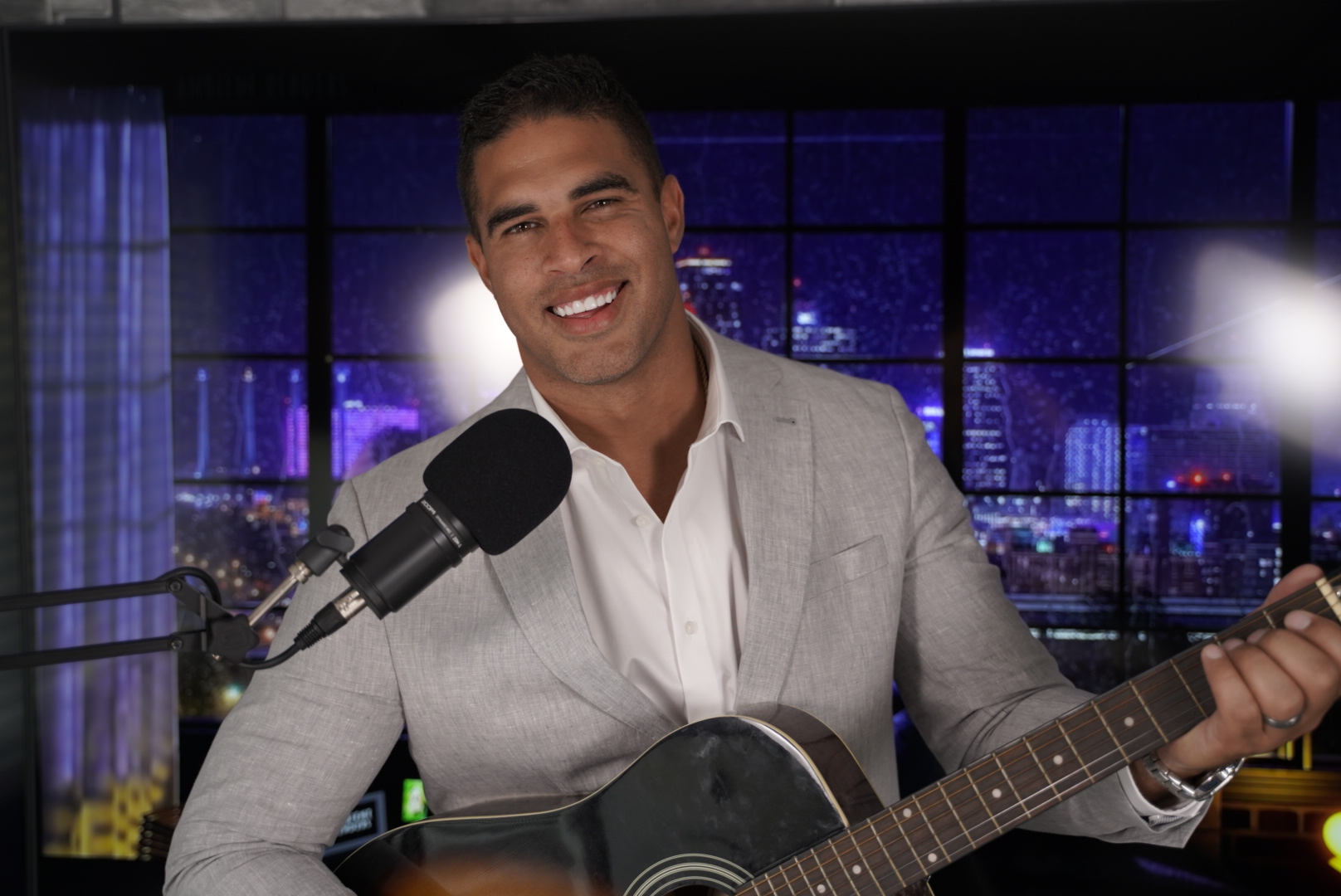 Brandon Mason in a gray blazer and white shirt holding an acoustic guitar beside a studio microphone